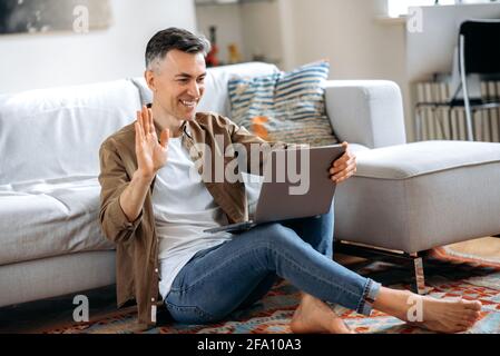 Handsome confident gray-haired caucasian senior man sitting in living room on carpet wearing casual clothes, using laptop, video chatting with friends or family, greeting with hand gesture and smiling Stock Photo