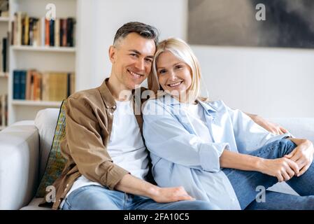 Portrait of a pleasant happy mature caucasian married couple relaxing on sofa in living room at home, spending time together, dressed in casual stylish clothes, smiling at camera, happy together Stock Photo