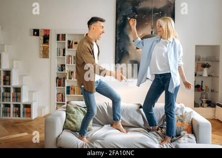 Happy caucasian senior married couple, crazy and funny spouses dancing on sofa in living room, enjoying success or victory, looking at each other, gesturing with hands, smiling, having fun Stock Photo