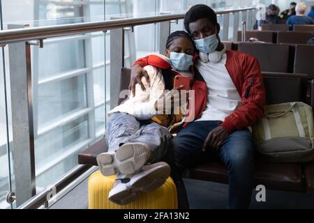 Couple of african tourists wear protective mask, sit together on chair in airport waiting for flight Stock Photo