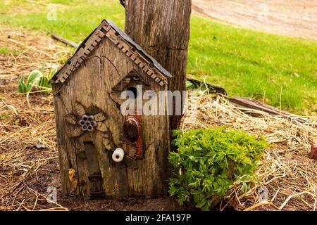 A rustic birdhouse made from a repurposed door handle, water faucet handle, belt, insulator, tin and old decorative wood leans against a post. Stock Photo