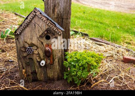 A rustic birdhouse made from a repurposed door handle, water faucet handle, belt, insulator, tin and old decorative wood leans against a post. Stock Photo