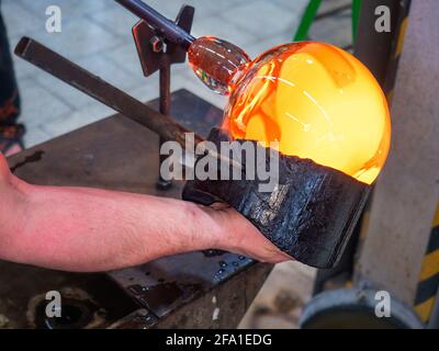 Glassblower is using wet wooden form for shaping the red melted glass on the end of glassblowing pipe. Traditional handmade Czech glass workshop in Ku Stock Photo