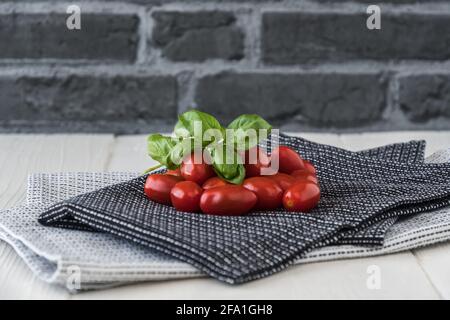 Ripe cherry tomatoes with basil branch on black and white tea towels against a gray stone background Stock Photo