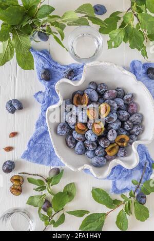 White curved bowl filled with ripe plums, some of them sliced, on a white wooden table with green leaves. Top view. Stock Photo