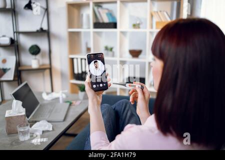 Concept of telemedicine and patient consulting online. Sick woman feeling bad at home, measuring body temperature, calling to male indian doctor using smartphon. Focus on phone screen Stock Photo