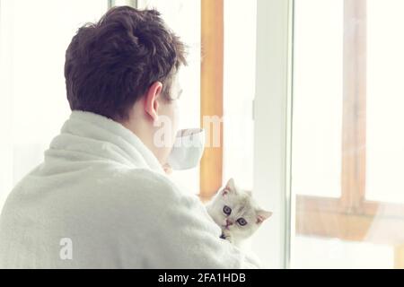 Young caucasian man is drinking coffee near the window. He is holding a cat. Early morning, sunshine. Stock Photo