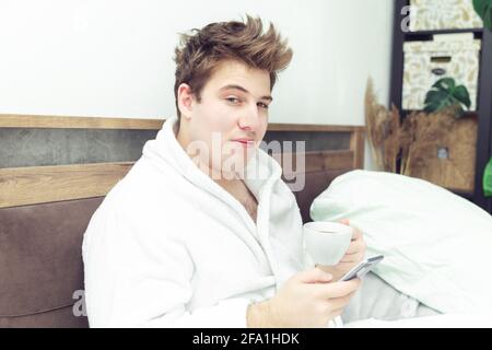 Young caucasian man in a bathrobe with a cup of coffee and a phone looking at camera. Early morning in bed. Stock Photo