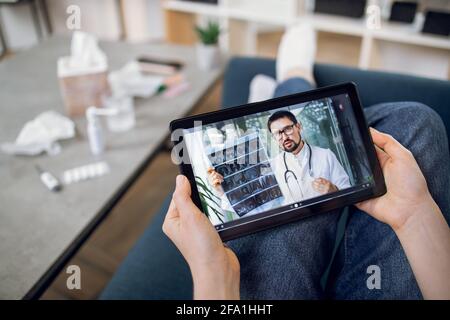 Unrecognizable female patient having video chat with male doctor on digital tablet at home, getting online consultation. Telemedicine and remote medical services. Stock Photo