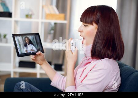 Young sick woman sits on couch at home and holds a spray for sore throat. Blur view of African female doctor talking to patient by video call on digital tablet. Online quarantine medical consultation. Stock Photo