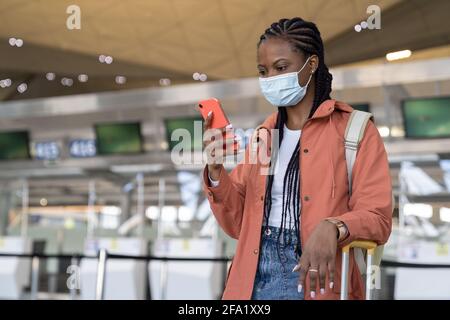 African woman with smartphone wear medical mask wait for flight in airport under covid-19 pandemic Stock Photo