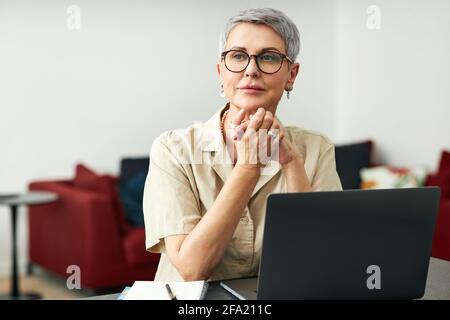 Stylish mature woman with grey hair wearing glasses sitting at home in front of a laptop Stock Photo
