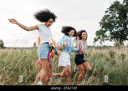 Four women holding hands running on field. Laughing friends having fun outdoors in summer. Stock Photo