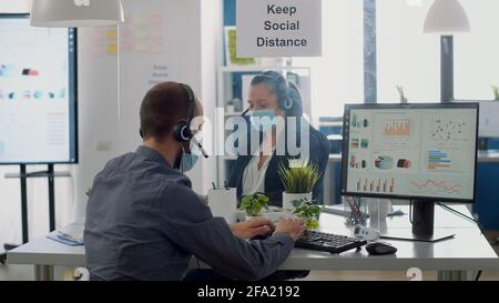 Collegues with protection medical face masks and headphones working in business office at management project during coronavirus pandemic. Coworkers keeping social distancing to prevent virus disease Stock Photo