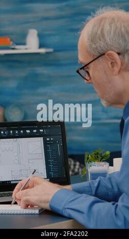 Senior man architect analysing digital prototype with plans from laptop working from home, taking notes on notebook. Elderly engineer designer using cad software to create a 3D concept of buildings. Stock Photo