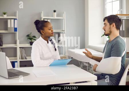 Millennial caucasian man patient with broken hand talk to afro american doctor Stock Photo