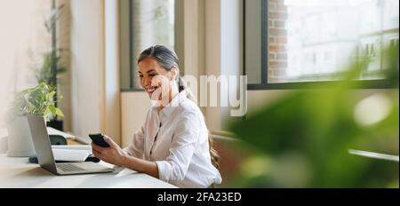 Mature woman sitting at office desk with mobile phone and laptop. businesswoman in office using phone. Stock Photo