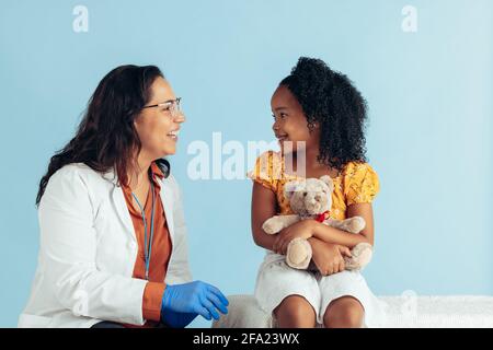 Doctor and girl smiling on blue background. Smiling pediatrician talking with cute patient in clinic. Stock Photo