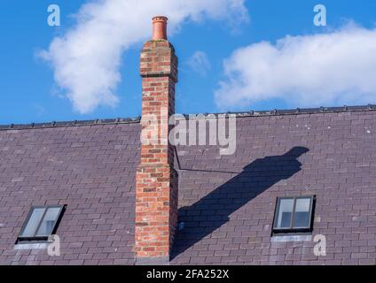 A tall red bricked chimney on a grey slate roof with blue sky background on a sunny day. Stock Photo
