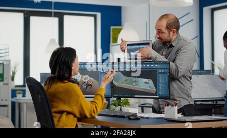 Team of architects and engineers working on building prototype project, using computers running 3D CAD Software. Project manager discussing with employee holding tablet explaining technical specifics Stock Photo