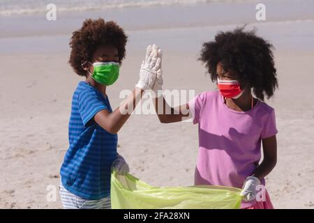 African american mother and daughter wearing face masks collecting rubbish high fiving at the beach Stock Photo