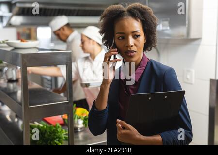 Mixed race kitchen manager talking on the phone with professional chefs in background Stock Photo