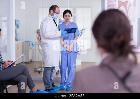 Dentist doctor holding teeth x-ray and nurse listening diagnosis in stomatology healthcare reception. People filling dentistiry form in waitting area. Stock Photo