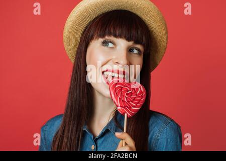 Sweet food, candy, confectionary and enjoying concept. Dreamy beautiful red haired woman in jeans dress and straw hat, licking colorful heart shaped lollipop isolated on red background