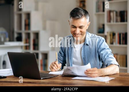 Satisfied successful confident mature gray-haired caucasian businessman, manager or freelancer, in casual stylish wear, working at office using laptop, checking work papers, taking notes, smiling Stock Photo