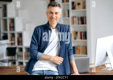 Portrait of a handsome confident successful mature gray-haired caucasian man, in casual stylish denim shirt, stands at the work desk, looks at the camera, smiles friendly Stock Photo