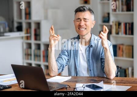 Mature gray-haired caucasian, male freelancer, top manager, in stylish clothes, works at a laptop, sits at work desk, worries, hoping for the desired result, gestures with his hands, closes his eyes Stock Photo