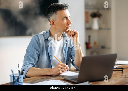 Pensive successful smart senior gray-haired Caucasian businessman, broker or freelancer in casual clothes, working remotely behind a laptop, planning a business project or report, looking to the side Stock Photo