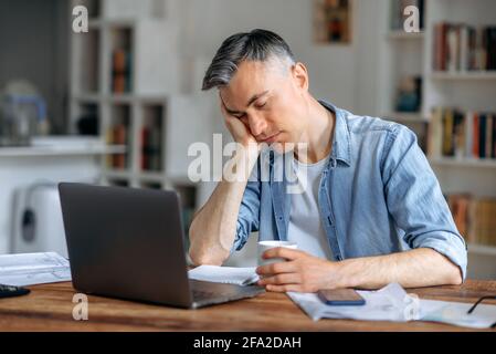 A sleepy tired attractive gray-haired senior manager, freelancer or office employee, in casual wear, fell asleep on his arm behind a laptop while work, took a break from work, need rest Stock Photo