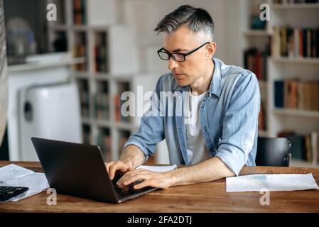 Focused smart successful gray-haired senior caucasian man wearing glasses, freelance, businessman or manager, works remotely on laptop in living room, texting with colleague or client,preparing report Stock Photo
