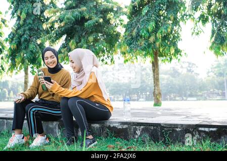 two girls in veil sits with chatting when exercise breaks while using handphone and drink water with bottle