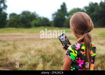 Defocus Selfie girl. Little girl takes a selfie by hand. Reflection on screen. Back view of preteen girl in summer standing on the meadow and taking a Stock Photo