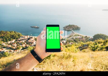 Man's hand holds a smartphone with a blank screen vertically on the background of Sveti Stefan island Montenegro Stock Photo