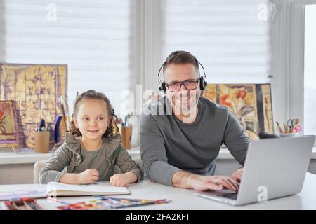 Cute small kid girl drawing next to dad who is working online in headphones with laptop at the table. Attractive family busy looking straight to camera happy smiling during covid-19 lockdown Stock Photo