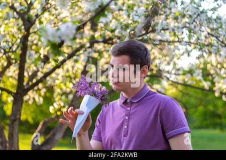 Defocus portrait of handsome caucasian man with cone flowers in purple shirt posing in lilac garden. Young brunette guy smelling fresh flowers and Stock Photo