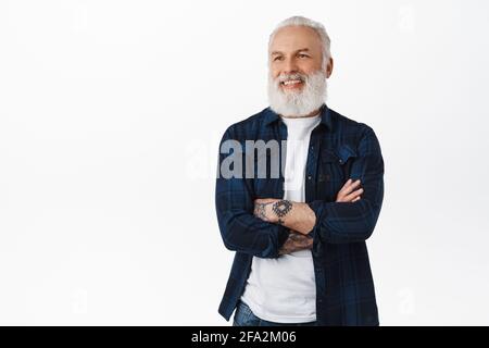 Smiling mature senior man with tattoos and grey long beard, cross arms on chest confident, looking left at promotional text logo, pleased to see Stock Photo