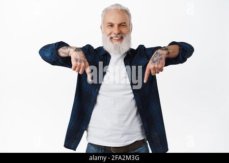 Looking down. Smiling handsome senior man, old hipster guy with tattoos pointing fingers bellow, showing advertisement on bottom with happy face Stock Photo