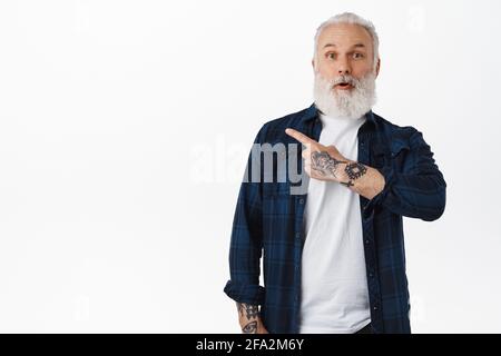 Surprised mature man with beard and tattoos, pointing finger left with amazed face, checking out cool advertisement, showing interesting promo offer Stock Photo