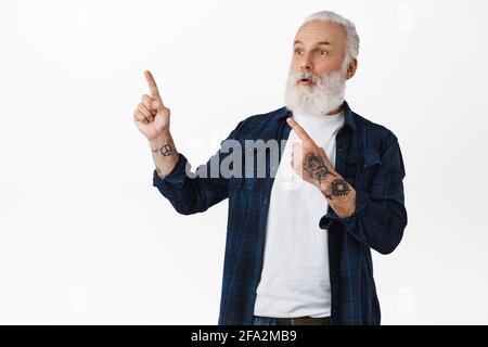 Surprised senior hipster guy pointing, looking at upper left corner, hands covered in tattoos, gasping amazed as checking out promo offer on top Stock Photo