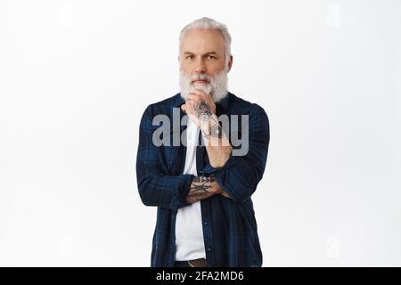 Thoughtful old man touching his beard and looking serious at camera. Senior guy with tattoos pondering decision, making choice, standing over white Stock Photo