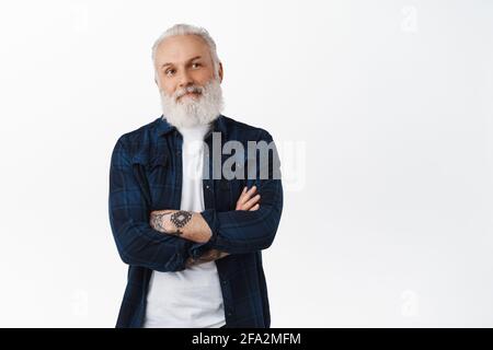 Smiling senior man with tattoos looking aside at promo logo, cross arms on chest and thinking, making decision, contemplating something on right side Stock Photo