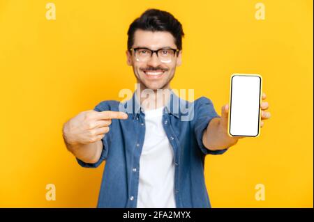 Defocused caucasian guy with glasses, looks at the camera, hold smartphone with white empty screen for your presentation in hand and points finger at it, stand on isolated orange background,copy space Stock Photo