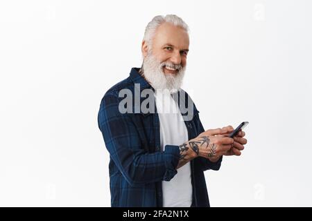Happy stylish senior man using mobile phone, messaging or browsing internet on smartphone and looking at camera with cheerful smile, standing over Stock Photo