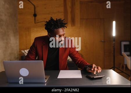 Handsome Black African American Man Working on Laptop Computer while Sitting Behind Desk in Cozy Living Room. Freelancer Working From Home Stock Photo