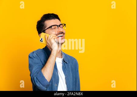 Pleasant phone conversation. Caucasian contented unshaven stylish guy with glasses is talking on the smartphone with friend or family, smiling, standing on isolated orange background Stock Photo