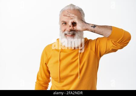 Happy old man with tattoos showing okay sign, looking through OK fingers and smiling pleased, satisfied, praise something good, say yes or alright Stock Photo